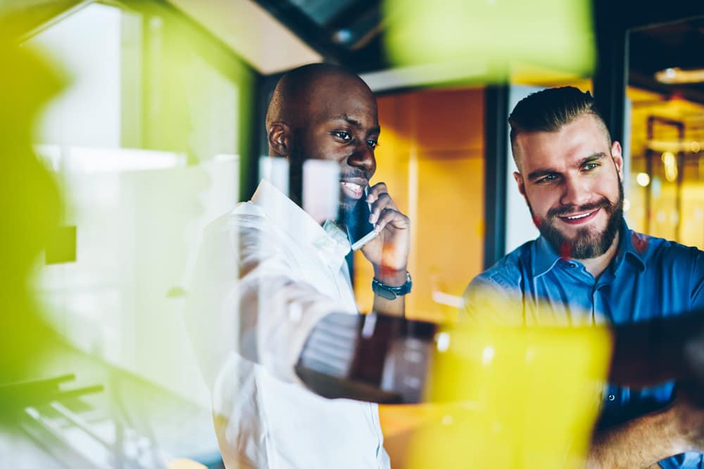 coworker calling on smartphone while discussing working plan together with colleague in office behind glass wall with sticky notes